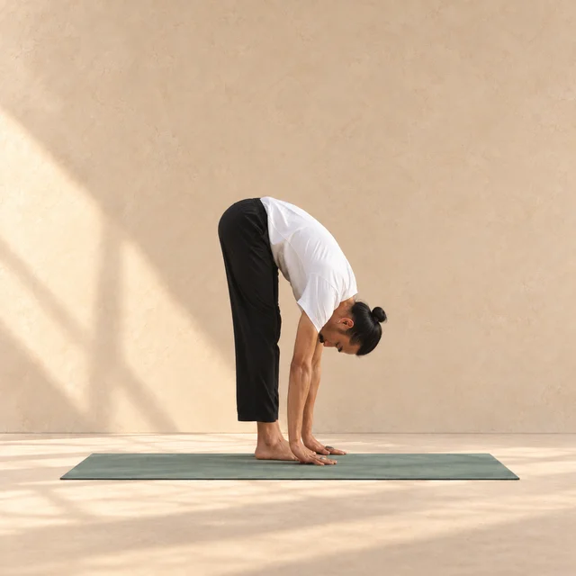 Uttanasana : modèle debout, buste plié vers les jambes et mains au sol sur un tapis sauge dans un studio beige.