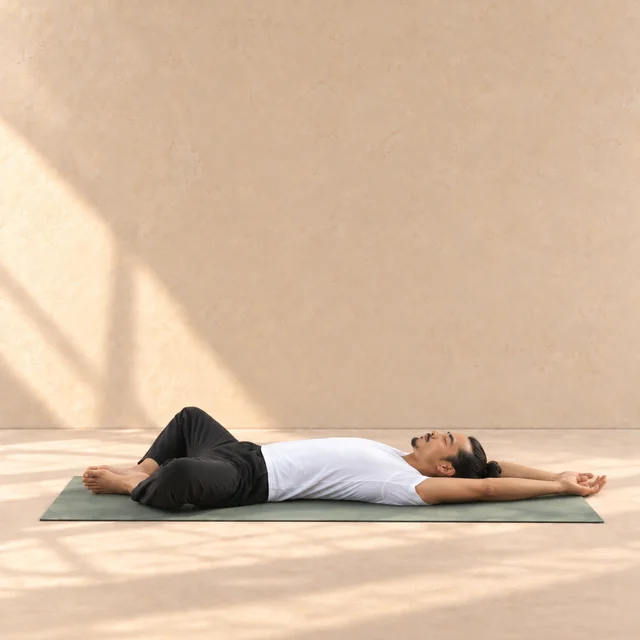 Supta Baddha Konasana de profil, dos au tapis, plantes de pieds ensemble, genoux ouverts et bras au-dessus de la tête.