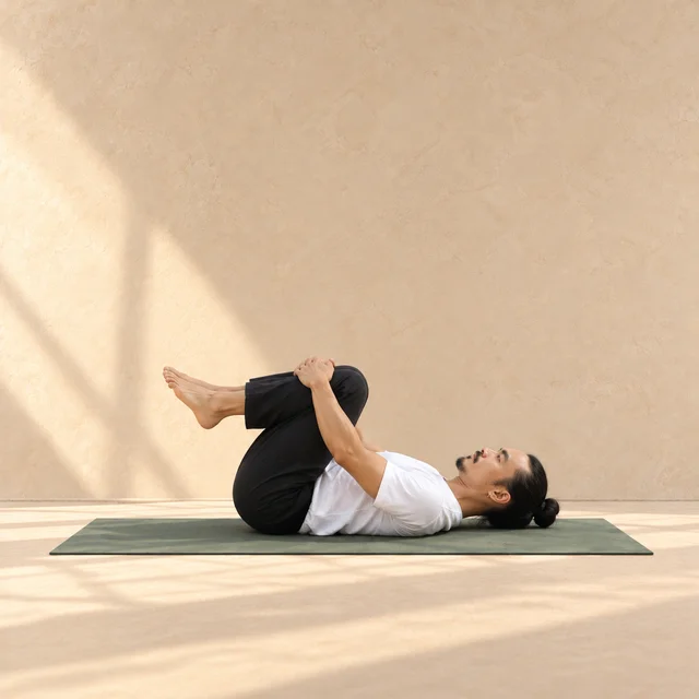 Apanasana de profil, tête à droite : homme allongé sur le dos, deux genoux pliés vers la poitrine, mains autour des tibias et épaules au tapis.